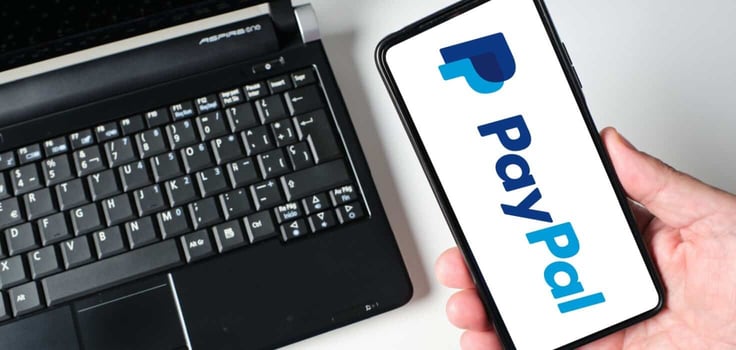 A person holds a smartphone displaying the Paypal payment application while sitting next to a laptop. This shows the integration of technology in financial transactions