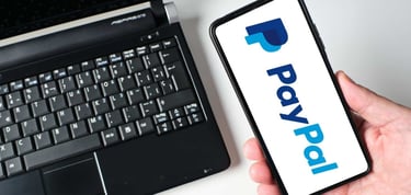 A person holds a smartphone displaying the Paypal payment application while sitting next to a laptop. This shows the integration of technology in financial transactions A person holds a smartphone displaying the Paypal payment application while sitting next to a laptop. This shows the integration of technology in financial transactions